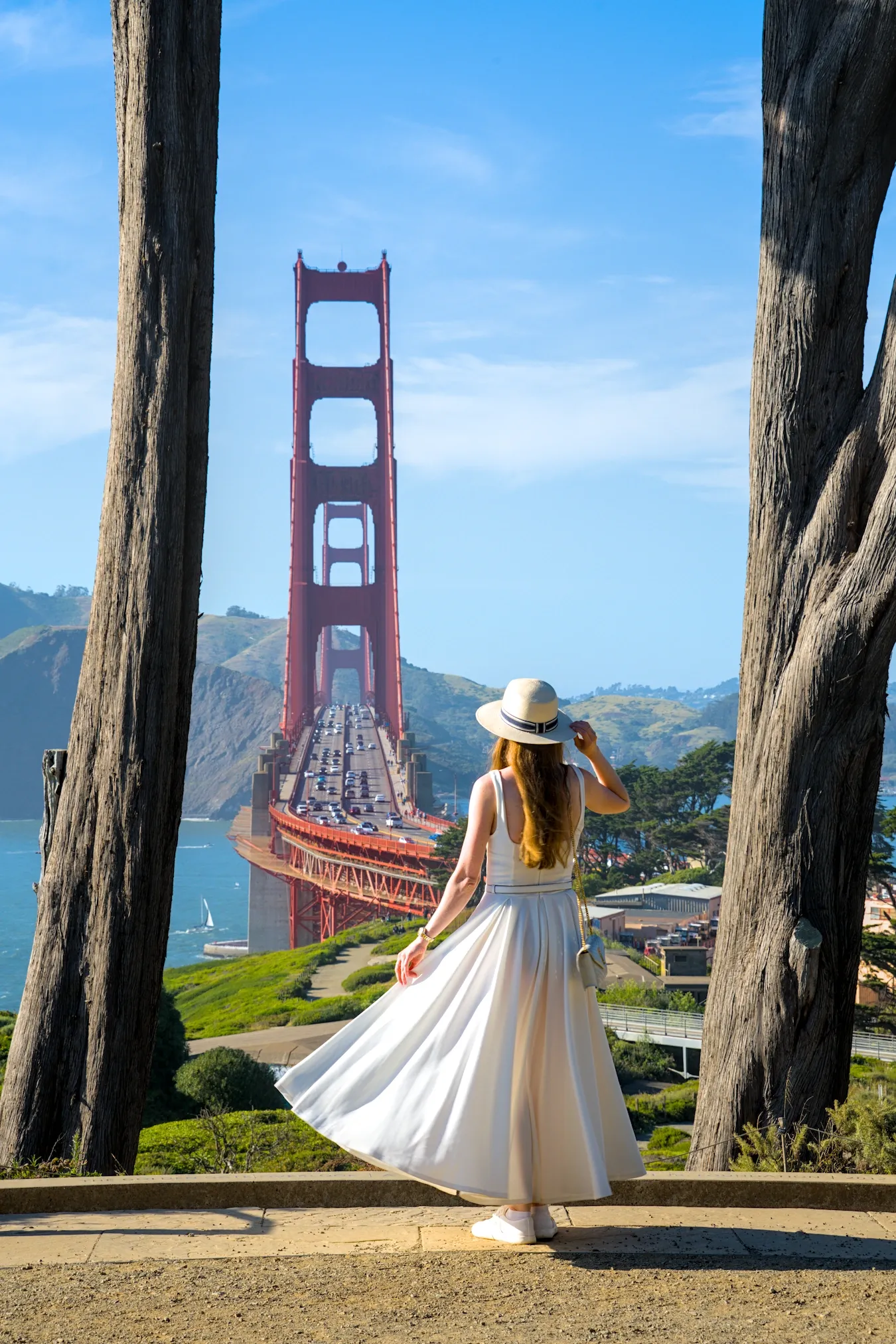 A woman in a white dress looking at the Golden Gate Bridge, framed by two cypress trees